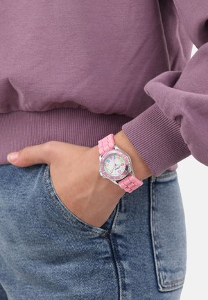 Rosa Gummiband-Uhr mit einem runden Ziffernblatt, das bunte Stundenmarkierungen aufweist. Silberner Rahmen und ein lässiges Denim-Outfit mit einem lila Pullover.