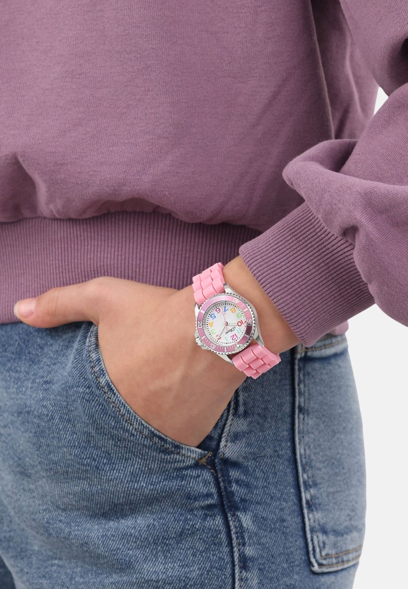 Rosa Gummiband-Uhr mit einem runden Ziffernblatt, das bunte Stundenmarkierungen aufweist. Silberner Rahmen und ein lässiges Denim-Outfit mit einem lila Pullover.