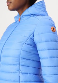 Světle modrá bundu z péřového materiálu s kapucí, s vodorovným švem, zipem vpředu a oranžovou nášivkou loga na levém rukávu.