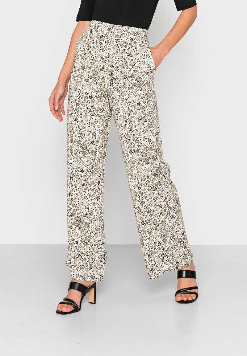 Pantalons larges floraux beige avec des accents verts et bruns, dotés d'une taille haute et de poches. Portés avec des sandales à talons noires.