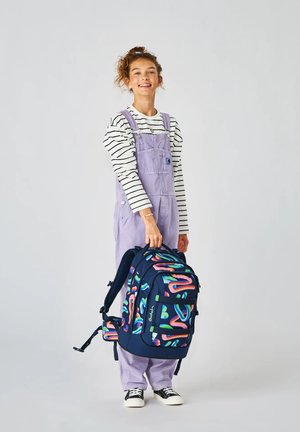 Lächendes Mädchen in lavendelfarbenen Latzhosen und gestreiftem Hemd, das einen großen dunkelblauen Rucksack mit bunten abstrakten Mustern hält.