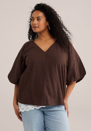 Žena s dlhými kučeravými vlasmi nosí hnedú blúzku s výstrihom do V a naberanými rukávmi a svetlomodré džínsy s čipkou, pózuje zboku.