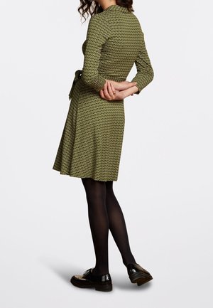 Zielona sukienka w wzory z rękawami o długości trzech ćwierć, z wiązanym paskiem w talii, rozkloszowanym dołem i noszona z czarnymi rajstopami i butami. Pokazany widok z tyłu.