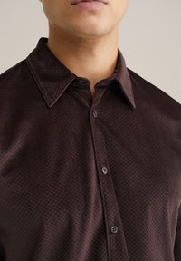 Camicia button-up marrone scuro con un motivo testurizzato, caratterizzata da un colletto classico e due bottoni visibili nella parte anteriore.
