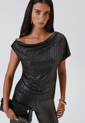 Vrouw in een zwarte glinsterende off-shoulder top en bijpassende broek, met een zwarte clutch en gouden armband en oorbellen.