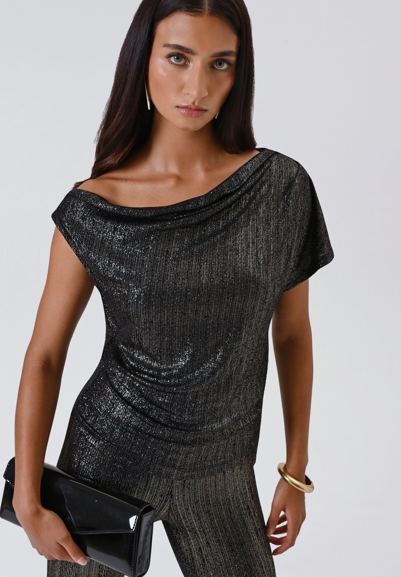 Donna che indossa un top nero luccicante senza spalline e pantaloni abbinati, tiene in mano una clutch nera e indossa un bracciale e orecchini dorati.