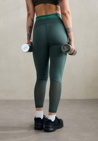 Zelené legíny s vysokým pasem a síťovinovými panely, s logem na pase a přiléhavým střihem. Modelka drží dva stříbrné činky.