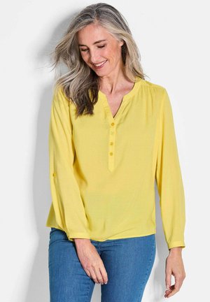 Blusa gialla leggera a maniche lunghe con scollo a V e dettagli con bottoni; texture morbida e vestibilità rilassata. Indossata con jeans blu.