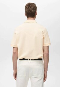 Polo tričko s krátkým rukávem z měkkého broskvového materiálu, s rovnoměrným střihem, decentním límečkem a kontrastními manžetami, nošené s bílými kalhotami.