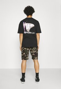 Svart t-shirt med en stor fjärilsdesign och "SHANGRI-LA" text på ryggen, tillsammans med kamouflagedesignade cargoshorts och svarta sneakers.