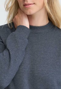 Grå genser med rund hals i strikket stoff, med lange ermer og ribbestrikkede mansjetter samt en glatt tekstur. Nærbilde av en modells utringning og arm.
