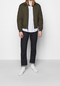 Osoba v zelené prošívané bundě, bílém vzorovaném tričku, tmavých vyhrnutých džínách, bílých ponožkách a bílých teniskách, stojící na pozadí jednoduché zdi.