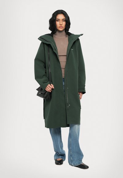 Manteau long vert avec fermeture éclair et fermeture à pression, doté d'une capuche, porté sur un top marron côtelé et un jean bleu évasé, avec un sac à main noir.