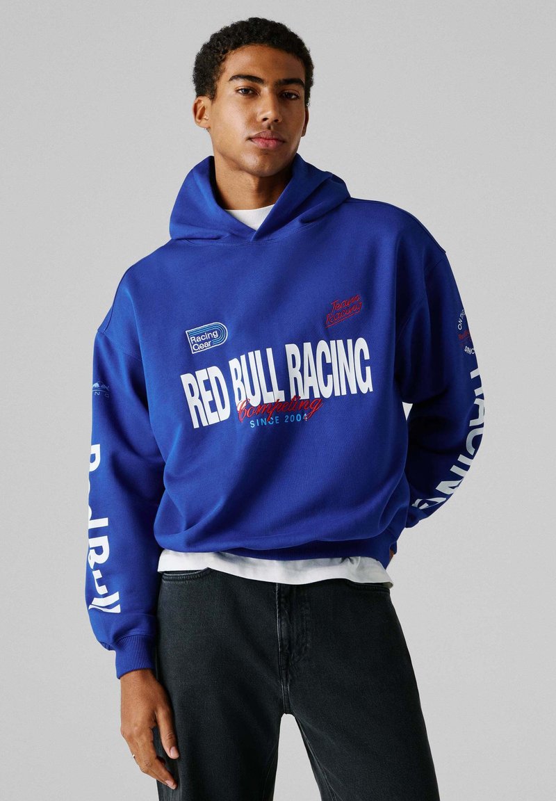 Νεαρός άνδρας που φοράει μπλε φούτερ Red Bull Racing με λευκό και κόκκινο κείμενο, στέκεται μπροστά σε απλό γκρι φόντο.