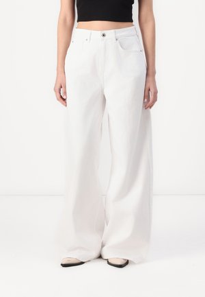 Osoba ubrana w białe, szerokie spodnie z wysokim stanem, zestawione z czarnym krótkim topem i beżowymi butami, stojąca na tle białej ściany.