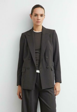 Frau mit dunklen Haaren, die einen dunklen zweireihigen Blazer und passende Hose trägt, steht vor einem schlichten hellen Hintergrund.