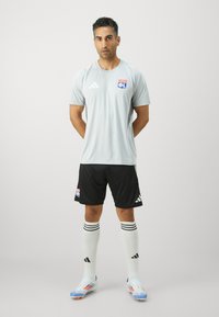 Lys grå adidas sportsskjorte med logo, svarte shorts, hvite sokker med svarte striper og fargerike fotballsko. Modellen står med armene bak ryggen.
