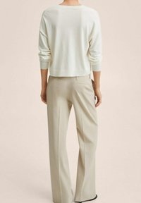 Person trägt ein weißes langärmeliges Oberteil und beige, weit geschnittene Hosen, steht mit dem Rücken zur Kamera vor einem schlichten Hintergrund.