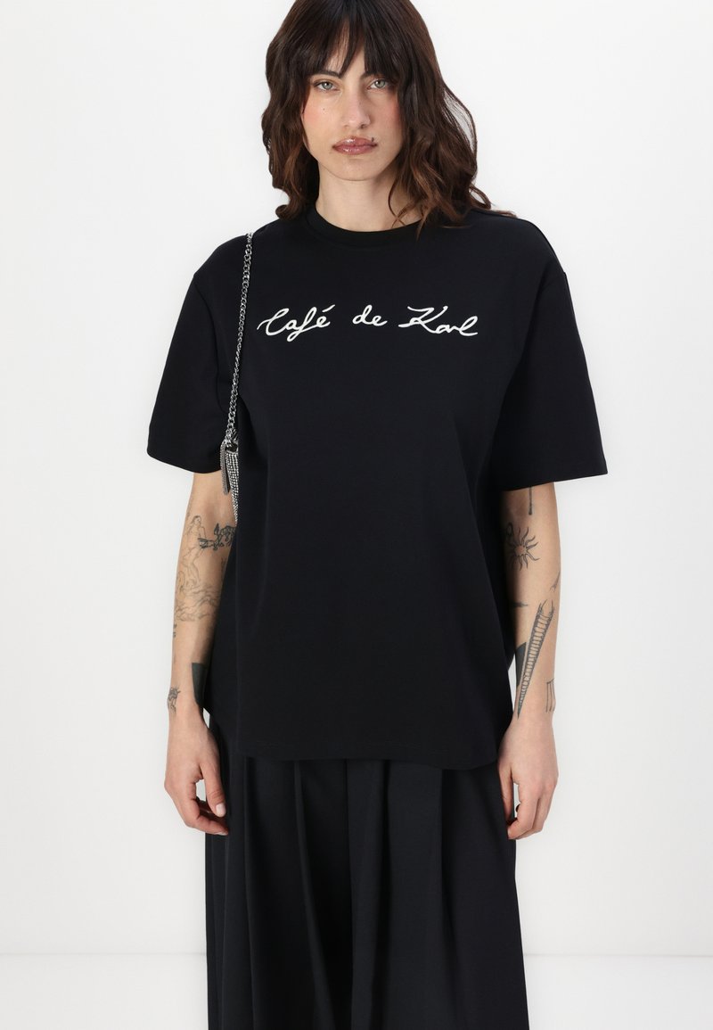 Schwarzes, übergroßes T-Shirt aus Baumwolle mit kurzen Ärmeln, das auf der Vorderseite einen weißen, geschwungenen Text „Café de Karl“ aufweist. Glatte Textur.
