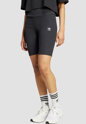 Čierne rebrované cyklistické šortky s bielym logom Adidas, v kombinácii s bielymi teniskami a prúžkovanými čierno-bielymi ponožkami. Jednoduchý dizajn.