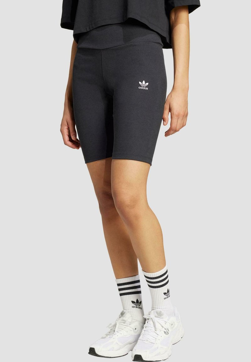 Svarta ribbade cykelshorts med en vit Adidas-logotyp, kombinerat med vita sneakers och randiga svartvita strumpor. Enkelt design.