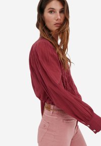 Blouse bordeaux à manches longues avec un tissu texturé et des broderies, associée à un pantalon rose clair ajusté et une fine ceinture en chaîne dorée.
