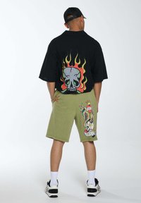 Maglietta nera oversize con un teschio colorato e fiamme stampate sul retro. Pantaloni corti verde oliva con una grafica dettagliata in stile tatuaggio.