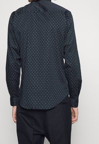 Tmavě modrá košile s dlouhými rukávy a zaobleným lemem, zdobená diamantovým vzorem v zlaté a bílé barvě. Vyrobeno z lehké látky.