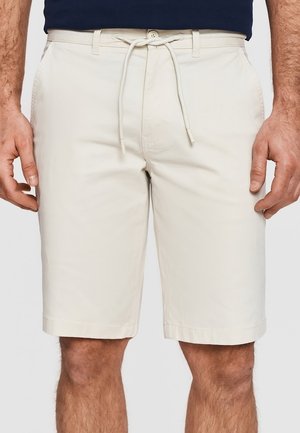 Beżowe krótkie spodenki męskie do kolan z przednimi kieszeniami, szlufkami na pasek i białym sznurkiem związanym w talii, noszone z ciemną koszulą.