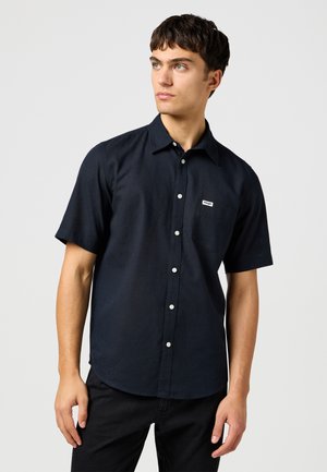 Nuori mies, jolla on yllään musta lyhythihainen nappipaita, jossa on pieni rintatasku, ja mustat housut, katsoo hieman vasemmalleen.