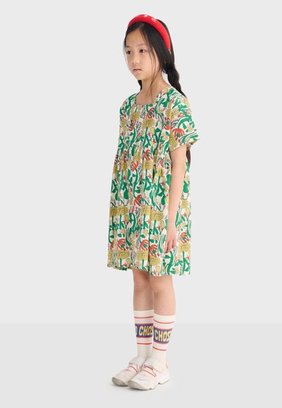 Jeune fille avec une longue tresse noire portant une robe à fleurs, des chaussettes blanches avec un texte bleu et jaune, des baskets blanches et un serre-tête rouge, debout de profil.