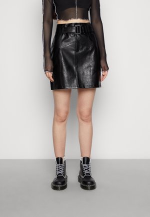 Personne portant une mini-jupe en cuir noire avec ceinture, un haut noir transparent à manches longues, et des bottes de combat noires à lacets, debout devant un fond uni.
