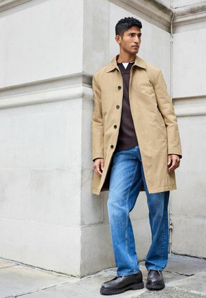 Trench beige con botones negros, suéter marrón, jeans azules y zapatos negros gruesos; de pie frente a una pared de piedra clara.