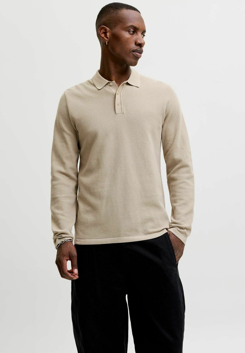 Homme portant un polo beige à manches longues et un pantalon noir, regardant sur le côté avec une main dans la poche et portant un bracelet chaîne.