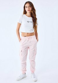 Weißes, kurz geschnittenes T-Shirt mit schwarzem Text, kombiniert mit hellrosa Cargo-Hosen mit mehreren Taschen und elastischen Bündchen, getragen mit weißen Sneakers.