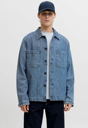 Veste en denim bleu clair avec un motif texturé, fermeture à boutons sur le devant, deux poches poitrine et un large col, portée sur une chemise grise.