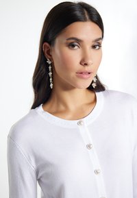 Cardigan bianco con bottoni a forma di cuore, caratterizzato da un collo rotondo e delicate cuciture. Orecchini pendenti con accenti di design floreale.