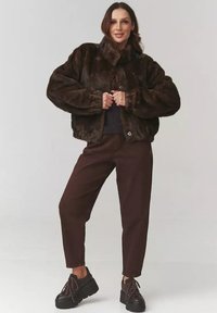 Brązowa kurtka z futra syntetycznego z wysokim kołnierzem, guzikami z przodu i obszernymi rękawami, w połączeniu z brązowymi spodniami o luźnym kroju i czarnymi masywnymi butami.