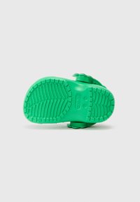 Chinelos Crocs verdes para crianças vistos pela sola, com aderência texturizada e tira ajustável para o calcanhar, em fundo branco simples.