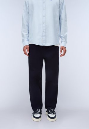 Mand iført lyseblå stribet skjorte med knapper, sorte lige bukser og sorte og hvide sneakers, stående mod en ensfarvet baggrund.