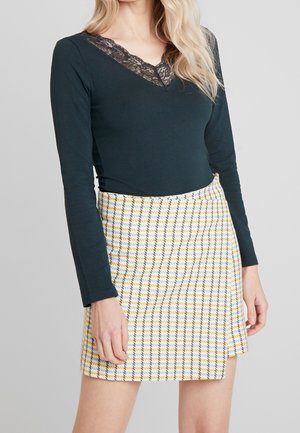 Mujer con una parte superior negra de manga larga con escote de encaje y una minifalda de cuadros amarilla y azul de talle alto.