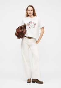 Γυναίκα με μακριά μαλλιά που φοράει λευκό τοπ κεντημένο με λουλούδια, λευκά παντελόνια φαρδιάς γραμμής, καφέ ζώνη, καφέ μπότες, κρατώντας μεγάλη καφέ δερμάτινη τσάντα.