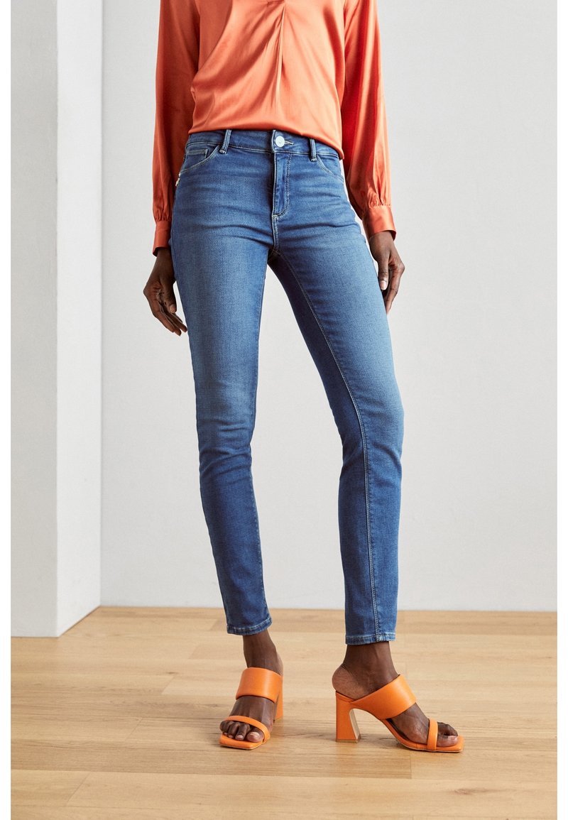 Personne portant un jean skinny bleu, un chemisier orange à manches longues et des sandales orange à talons hauts et bouts ouverts, debout sur un sol en bois.