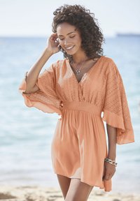 Perzikgekleurde kaftanjurk met een v-hals, klokmouwen, gestructureerde stof en een gegatherde taille; gedragen voor een strandachtergrond.