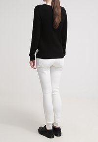 Czarny dzianinowy sweter z okrągłym dekoltem, ozdobiony akcentem lamówki z tyłu. Połączony z białymi obcisłymi jeansami i czarnymi butami.