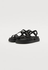 Zwarte sandalen hebben meerdere overlappende banden, een vlakke zool en een gladde textuur. Verstelbare gesp aan de zijkant voor een veilige pasvorm.