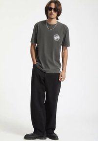 Donkergrijs katoenen T-shirt met een rond logo gedrukt op de linkerborst, gecombineerd met loszittende zwarte broek en zwarte schoenen.