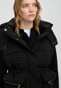 Czarna, ocieplana kamizelka o fakturowym, marszczonym wzorze. Posiada wysoki kołnierz, kieszenie na zamek błyskawiczny oraz zapięcia na guziki na ramionach. Podszewka wewnętrzna.