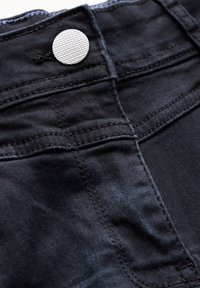 Jeans in denim nero con un bottone metallico testurizzato, cuciture visibili e una finitura liscia, che evidenziano un design aderente e un aspetto elegante.