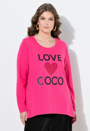 Kvinde med bølget hår har store hængeøreringe på og en lys pink langærmet skjorte med teksten "LOVE" og "COCO" samt et hjerte grafik.